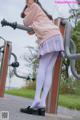 A woman in a pink sweater and plaid skirt leaning against a metal pole.