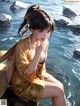 A woman in a yellow kimono sitting on a rock by the water.